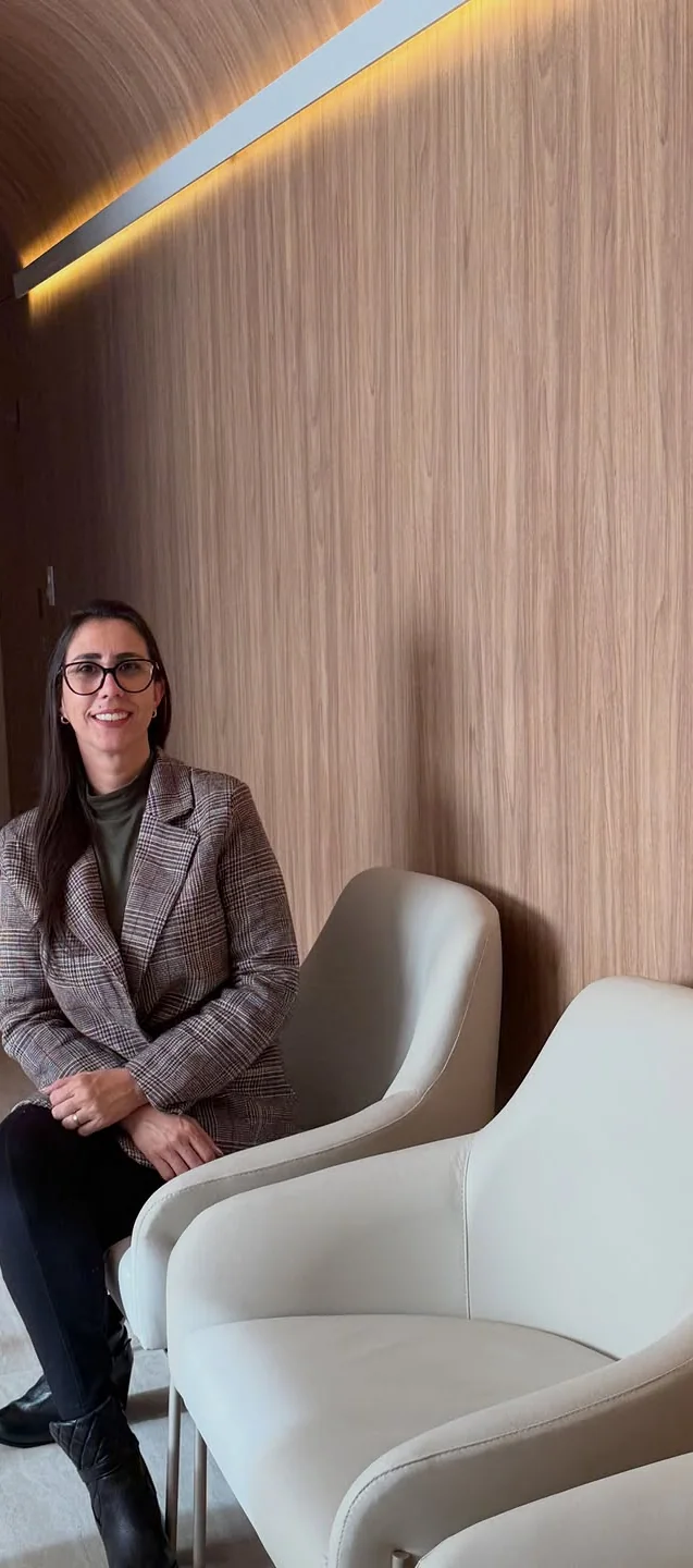 Débora sentada numa poltrona clara na sala de espera da Clínica Allmare, com paredes de madeira